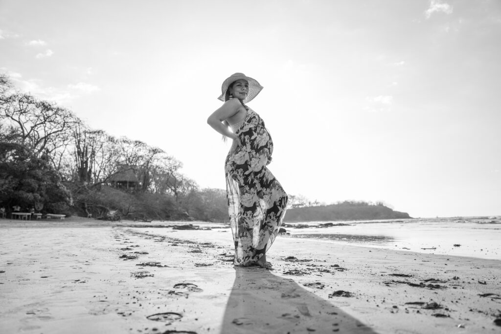 Black and white maternity portrait photography in Tamarindo, Costa Rica. A pregnant woman from the side, wearing a dress and a big hat, at the beach.