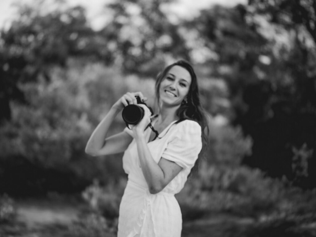 Black and white Portrait of Milena Re with camera, smiling, in a white dress, in the middle of plants