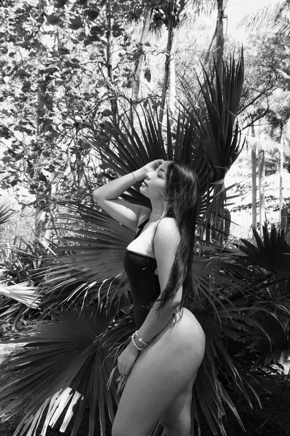 Black and white portrait photo of a young woman with long hair, in a swimsuit, holding a hand on a side of her face. In front of a plant.