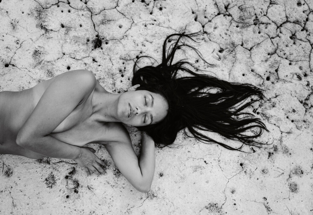 Black and white portrait photo of a young woman with long, black hair, laying on a dry ground