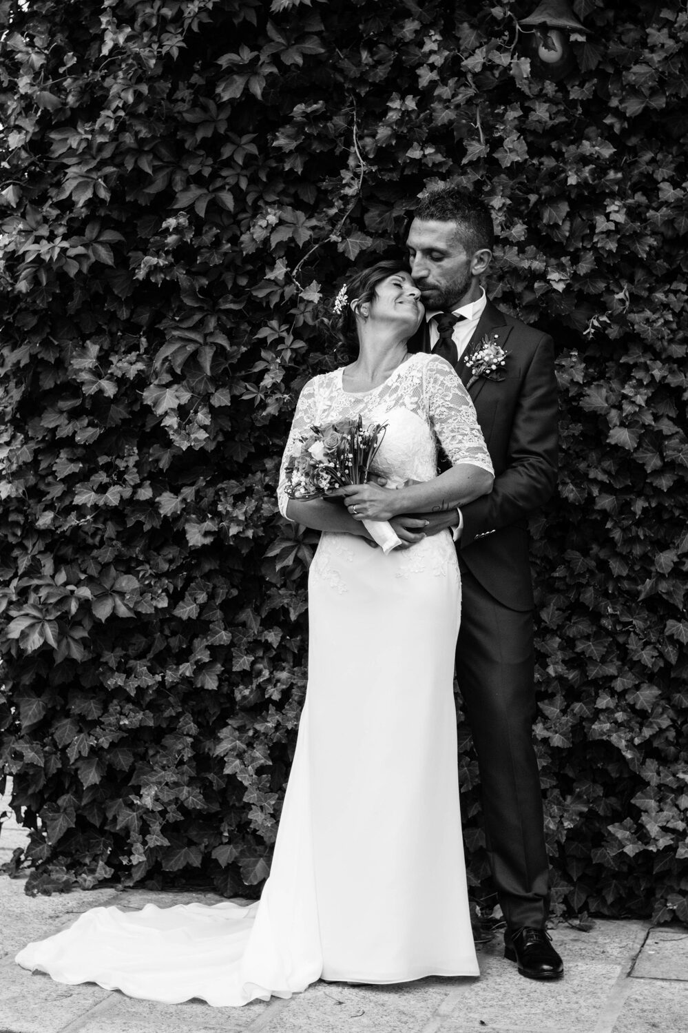 Black and white wedding photo of husband and wife hugging in front of a plant-wall