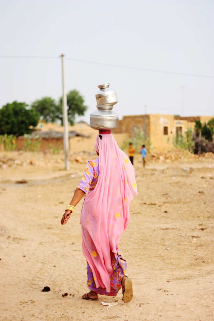Travel photo of a woman wearing a long pink headscarf, and caring silver vases on her head, in India.